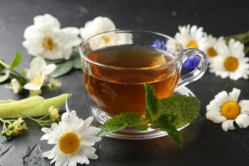 Aromatic herbal tea in glass cup, different flowers and mint on black table, closeup