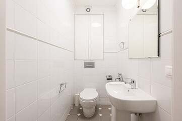tidy, small bathroom with white tiled walls, a toilet, a sink, and a mirrored cabinet. The floor...