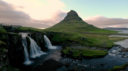 Breathtaking view of Kirkjufell mountain surrounded by vibrant greenery and cascading waterfalls at dusk in Iceland. Experience the serene beauty of Icelandic nature at its finest.