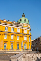 Bright yellow historical building in Rijeka, Croatia, with a domed green roof, classical windows...