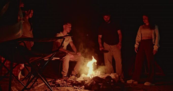 Friends gather around a bonfire at night, sharing stories and laughter under the starry sky. The warm glow of the fire creates a cozy and inviting atmosphere.
