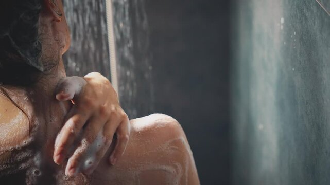 close-up hand strokes touches cleanses shoulder skin washes away white soap foam gel. happy woman taking shower rain under water drops. Girl enjoying bathing back rear view wet hair healthy self care