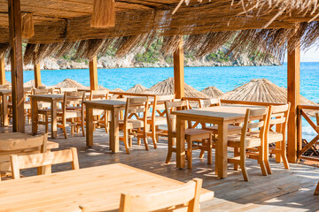 A charming coastal dining area features wooden tables and sun umbrellas, providing a perfect view of the clear blue sea.