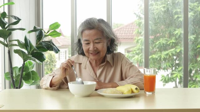 Healthy elderly Asian woman sits down to eat lunch with meatless, easily digestible congee and a banana and freshly squeezed orange juice rich in vitamin C with satisfaction.