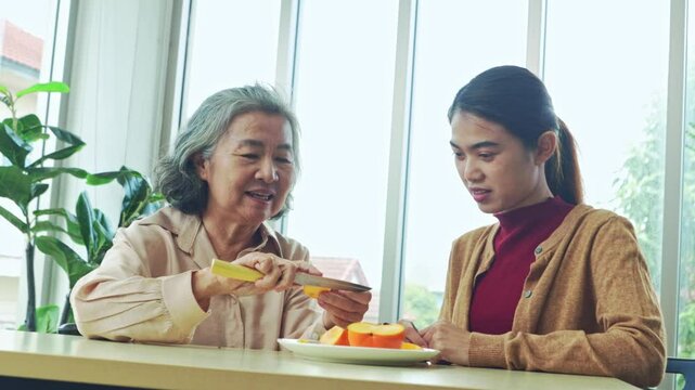 Loving moment between an elderly mother and her adult daughter eating healthy fruits. The elderly mother uses knife to peel persimmons for her daughter eat happily.
