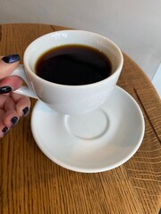 a white cup of coffee with a white saucer held by a woman's hand on a wooden table