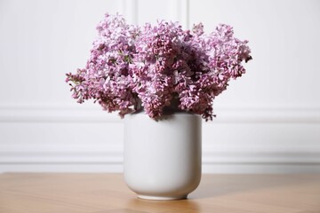 Beautiful lilac flowers in vase on wooden table near white wall indoors