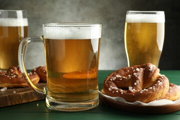 Tasty pretzels and glasses of beer on green wooden table against grey background, closeup