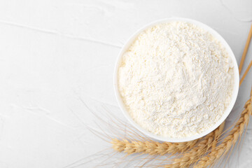 Wheat flour in bowl and spikes on white table, flat lay. Space for text