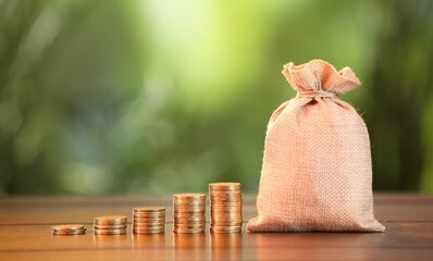 Burlap bag and stacked coins on wooden table against blurred background