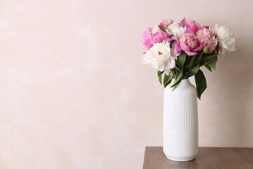 Bouquet of beautiful peonies in vase on wooden table near beige wall indoors, space for text