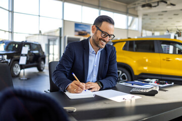 Male car salesman in a formal suit sitting at a desk in a bright, modern dealership showroom,...