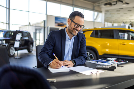 Male car salesman in a formal suit sitting at a desk in a bright, modern dealership showroom, signing a contract and preparing to finalize the sale of a new car to a customer.