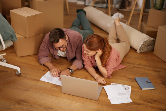 A couple is lying on the floor of their new home, looking at a laptop together. Unpacked boxes surround them as they plan their move, discussing important details and paperwork.