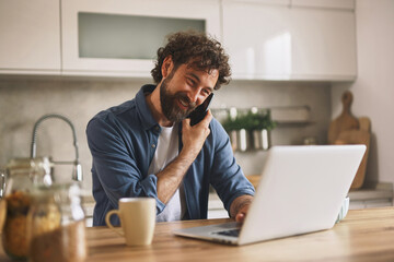 Man smiles while talking on the phone and using a laptop at a modern kitchen table, enjoying a warm beverage in bright daylight during his home office hours.