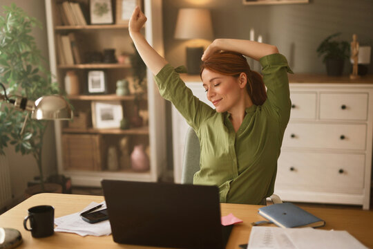 A woman in a green shirt takes a moment to stretch and relax in her home office. Sunlight filters through the window, creating a peaceful workspace ambiance filled with stationery and plants.