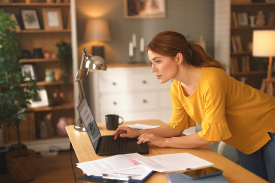 A woman leans forward, focused on her laptop, surrounded by papers and a coffee cup in a warm, inviting home office filled with books and decor.