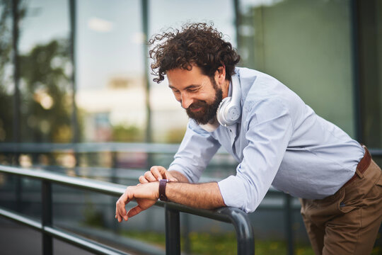 A man stands casually leaning on a railing in a contemporary outdoor space, smiling as he listens to music through his headphones, embodying a relaxed business vibe.