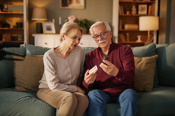 An elderly man and woman sit on a couch in their home, reviewing prescription medication, reading instructions on the label, and confirming dosages of pills during the daytime.