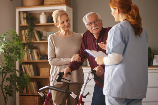 A home healthcare nurse in scrubs holds a clipboard and speaks with an elderly man, who is using a walker, and his wife in their living room, during a home care visit. - Powered by Adobe