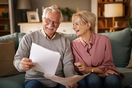 An elderly couple sits on a blue sofa in their living room, happily looking at papers. They smile at each other, seemingly pleased with what they are reading.