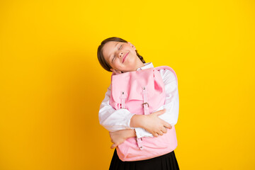 Adorable schoolgirl hugging her pink backpack with a cheerful face, isolated on a vibrant yellow...