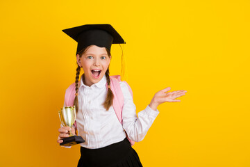 Excited young schoolgirl with graduation cap and trophy on vibrant yellow background celebrating...