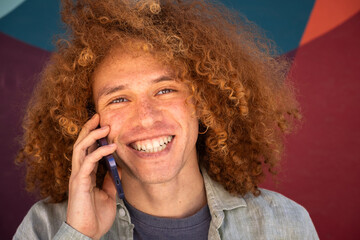 Close-up portrait of smiling young man with curly hair talking on smartphone against colorful wall. Cheerful student, modern communication, youthful lifestyle and personal branding concept.