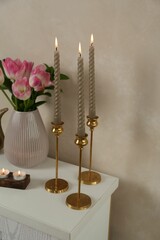 Elegant candlesticks with burning candles and tulips in vase on dresser near beige wall