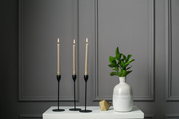 Elegant candlesticks with burning candles, branches in vase and other decor on dresser near grey...