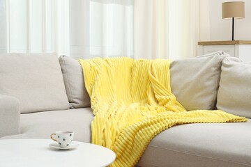 Comfortable sofa with yellow woolen blanket and cup on table indoors