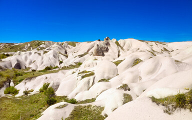 Amazing limestone eroded rock formations under clear blue sky in Cappadocia, Turkey. Unique terrain of Cappadocia shaped by wind and water, covered with sparse green vegetation and patches of grass.