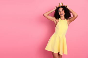 Young woman posing in a yellow dress with a playful crown styled fashion against a bright pink...
