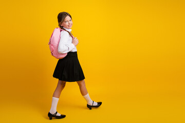 Young girl wearing school uniform carrying backpack smiling confidently against vibrant yellow...