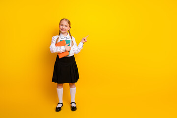 Smiling schoolgirl in uniform holding book on yellow background pointing with her finger towards...
