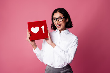 Young woman holding a red box with a heart symbol against pink background embodying happiness and...