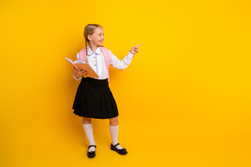 Happy schoolgirl with book wearing uniform pointing on a yellow background showing academic...