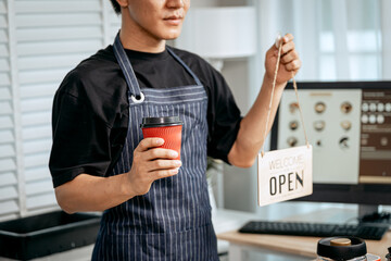 A young Asian male barista, dressed in a striped apron, skillfully creates beautiful latte art in a cozy, modern cafe, takeaway cups. holding open sign