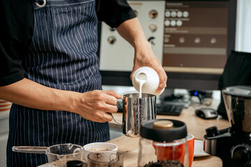 A young Asian male barista, dressed in a striped apron, skillfully creates beautiful latte art in a cozy, modern cafe, professional coffee equipment and takeaway cups.