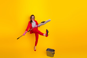 Stylish young woman in a vibrant red suit posing energetically with a mop on a bright yellow background