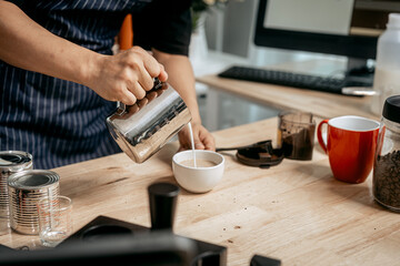 A young Asian male barista, dressed in a striped apron, skillfully creates beautiful latte art in a cozy, modern cafe, professional coffee equipment and takeaway cups.