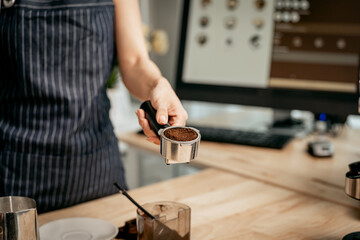 A close-up of a barista’s hand holding a portafilter filled with freshly ground coffee, ready for brewing. The striped apron in background suggests a professional cafe and expert preparation.