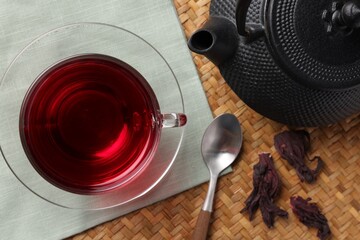 Delicious hibiscus tea in glass cup, teapot, dry roselle sepals and spoon on wicker mat, flat lay