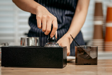 A young Asian female barista in a striped apron carefully pours steamed milk to create intricate...