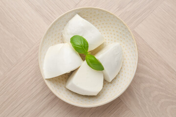 Pieces of delicious mozzarella cheese and basil in bowl on wooden table, top view