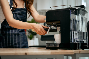 A young Asian female barista in a striped apron carefully pours steamed milk to create intricate latte art, working in a cozy modern café with professional coffee equipment and takeaway cups.