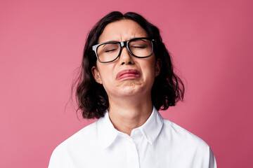 Expressive young woman in glasses wearing a white shirt displaying emotions against a pink...