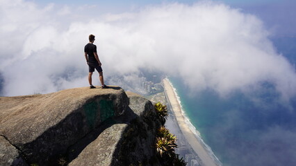 Epic aerial view from Pedra da Gávea in Rio de Janeiro, showcasing dramatic cliffs, lush rainforest, and the stunning coastline merging with clouds and the Atlantic Ocean