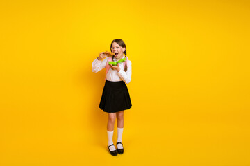 Smiling schoolgirl in uniform eating lunch, posed on yellow background showcasing learning and...
