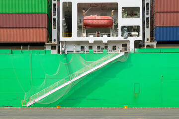 A vibrant green cargo ship docked, showcasing its access gangway, safety net, and a red life raft....